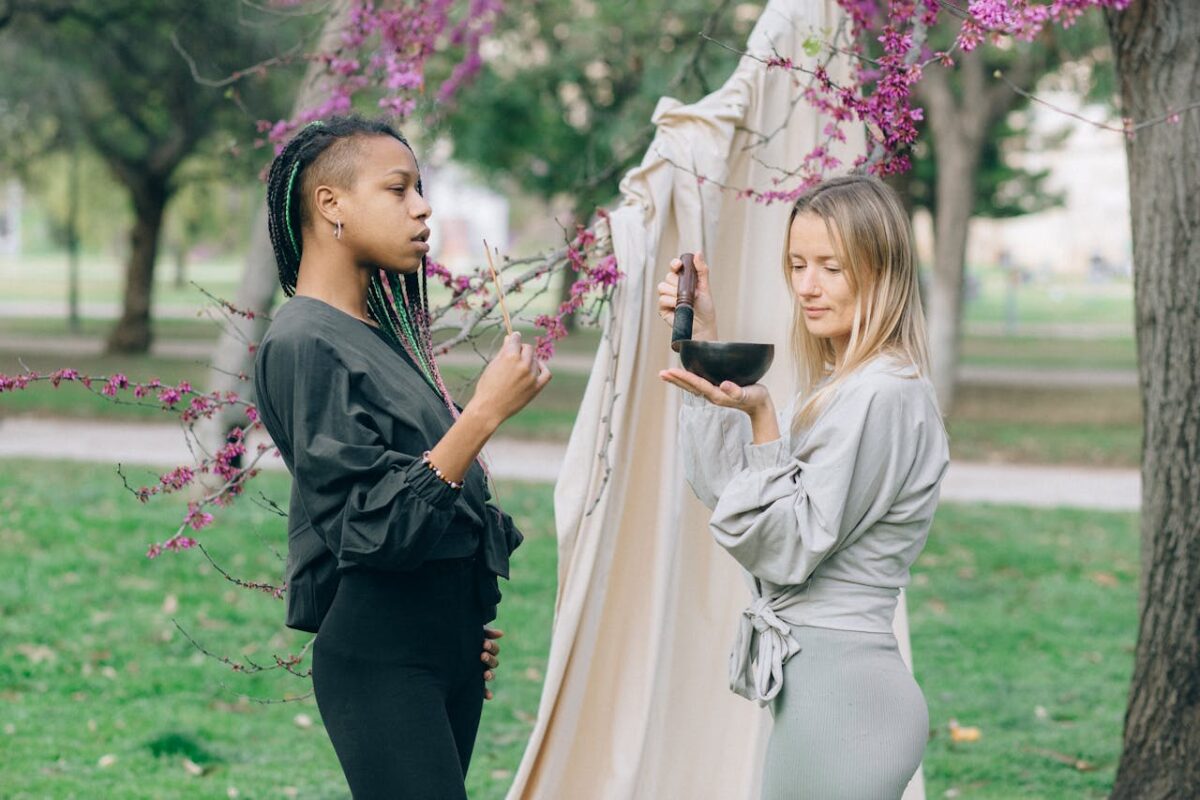 Two women engaging in a mindful practice outdoors, using a singing bowl near blooming trees.